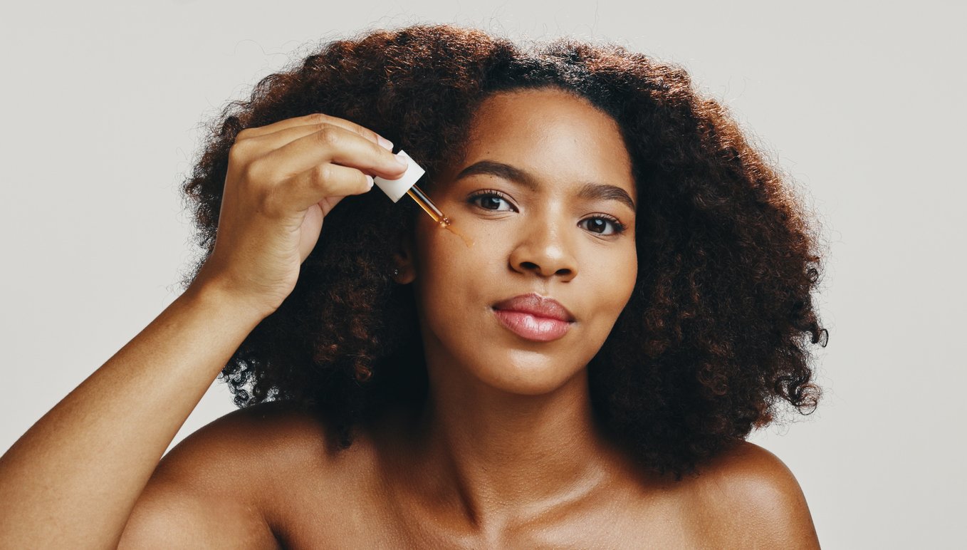 Closeup einer jungen Frau mit dunklem Hautton, die mit einer Pipette Serum auf ihre Wange gibt und in die Kamera schaut